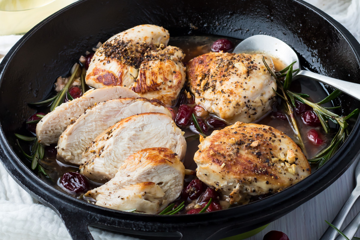 Cooked cranberry chicken breasts, some whole and some sliced, in a cast iron skillet with herbs and cranberries served alongside a metal spoon.