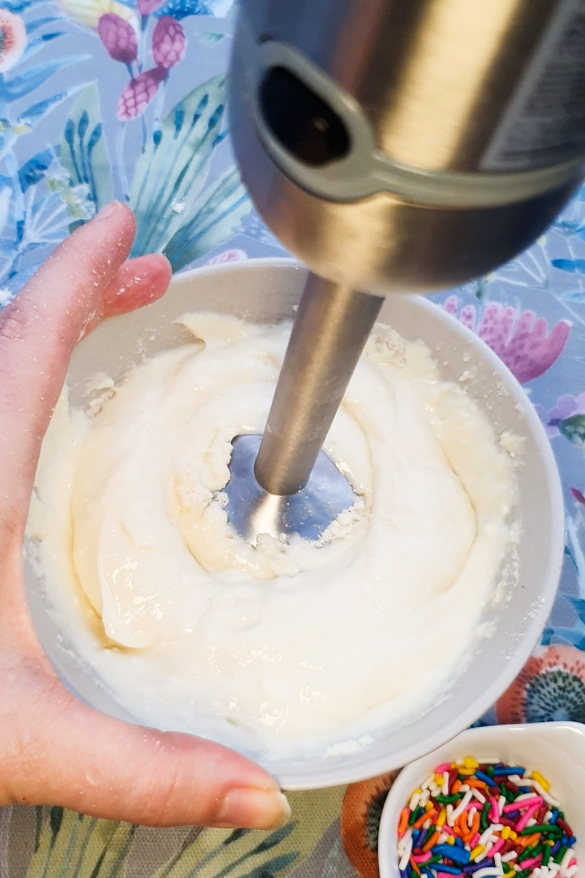 A hand holds a bowl of thick white mixture being blended with an immersion blender. Colorful sprinkles sit in a small dish nearby.