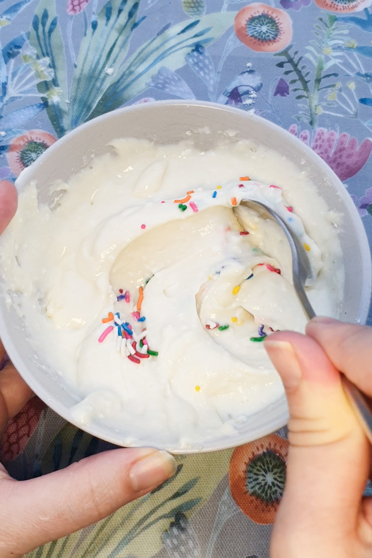 A person stirs white dip with rainbow sprinkles in a white bowl using a spoon, on a floral-patterned surface.
