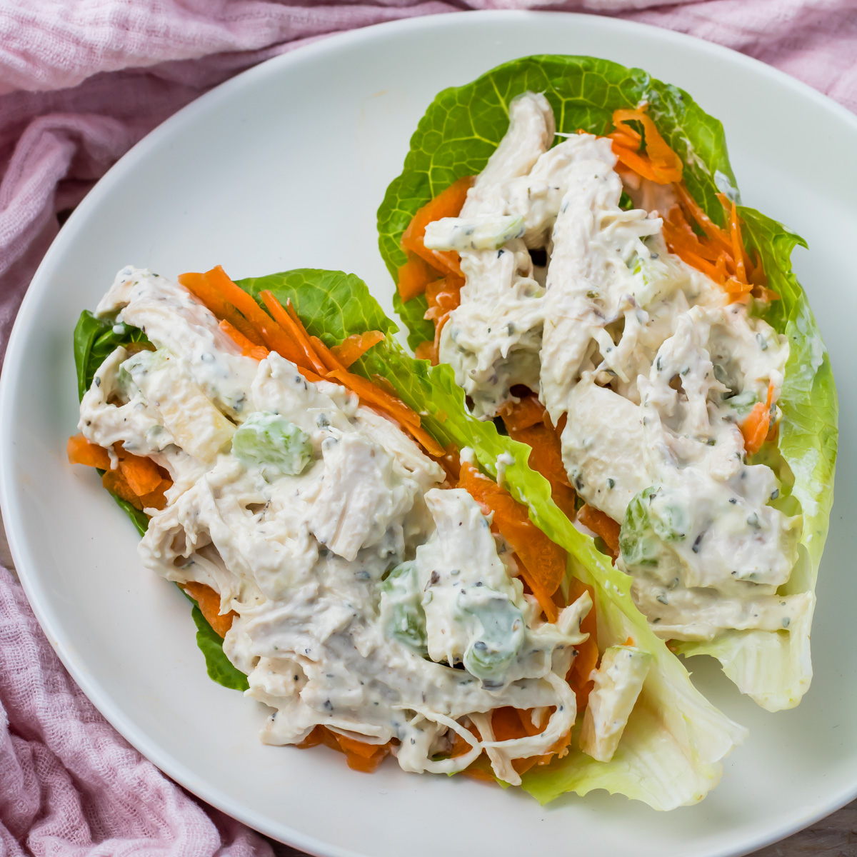 High protein chicken salad lettuce wraps with celery and carrots, served as lettuce wraps on romaine leaves atop a white plate, set off by a pink cloth underneath.