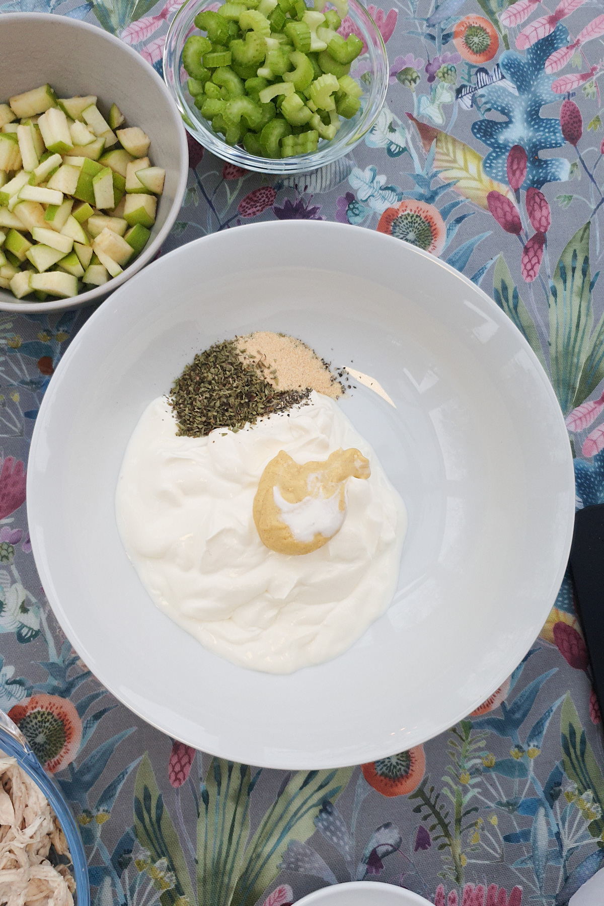 A white bowl with greek yogurt, mustard, dried herbs, and seasonings sits on a floral tablecloth. Nearby bowls contain chopped celery, green apple, and shredded chicken.