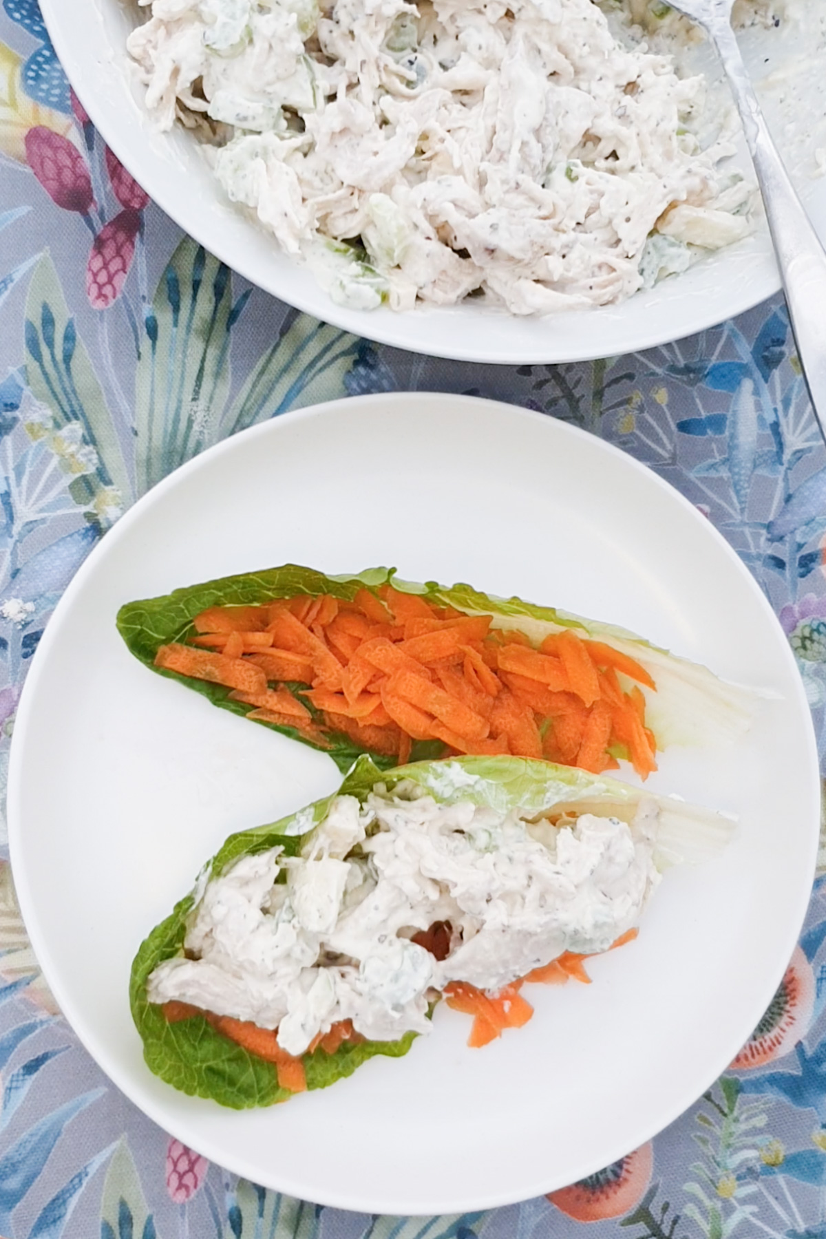 Two lettuce leaves on a white plate topped with shredded carrots and creamy chicken salad, with a bowl of the salad in the background on a floral tablecloth.