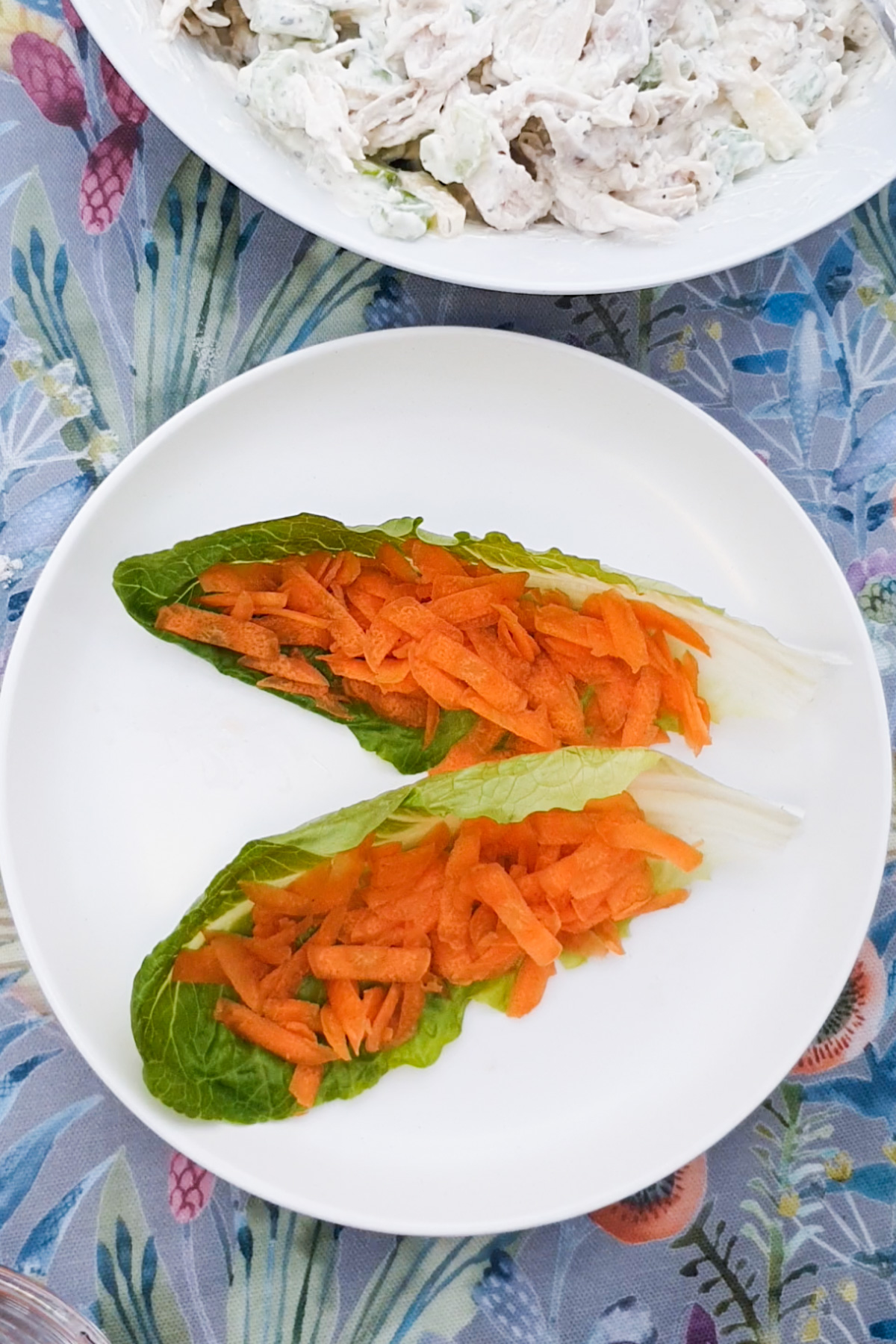 Two romaine lettuce leaves topped with shredded carrots attractively arranged on a white plate set atop a floral-patterned tablecloth.