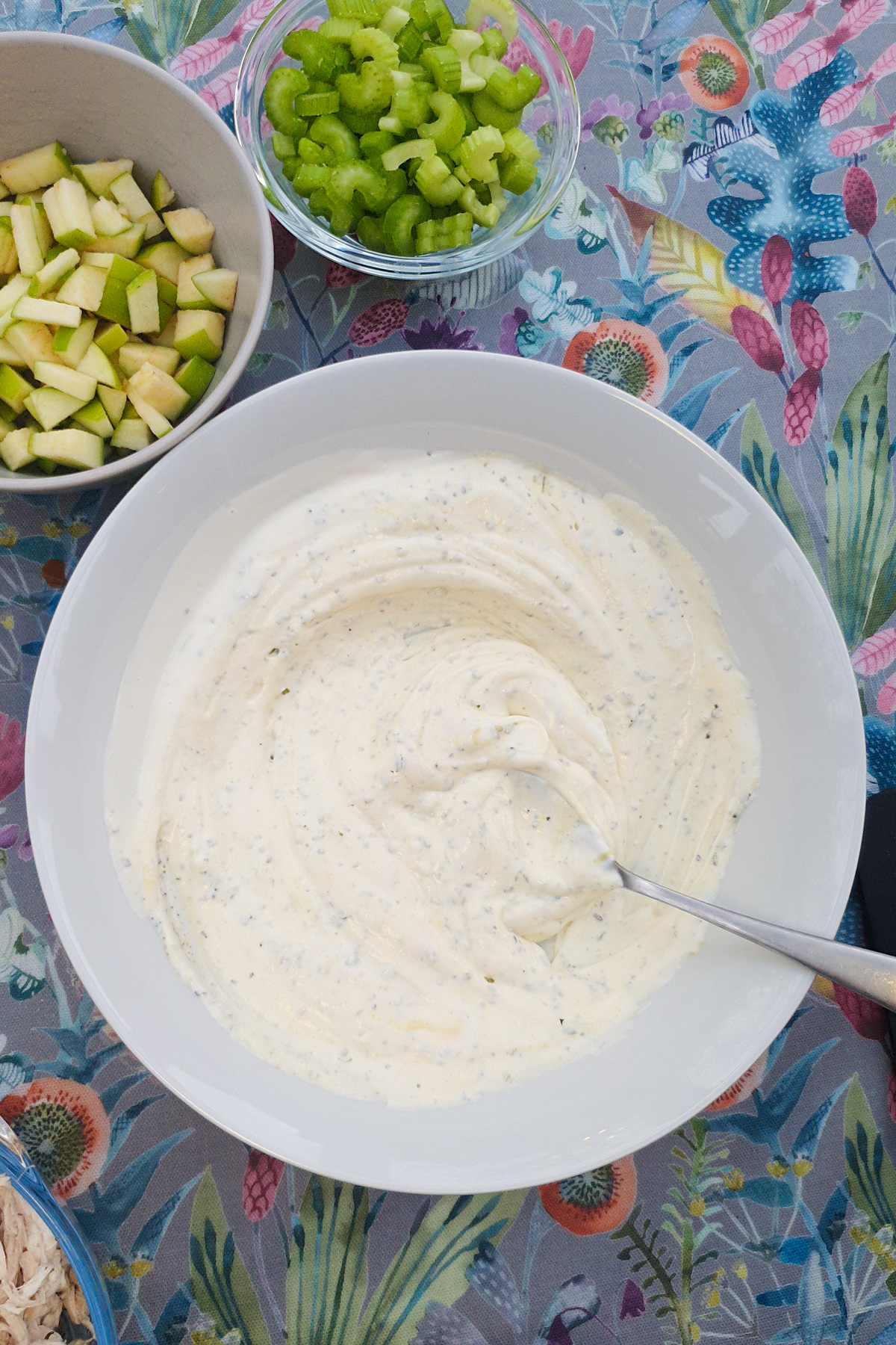 A bowl of creamy white dressing with herbs and a spoon sits beside bowls of chopped celery and green apple on a floral tablecloth.