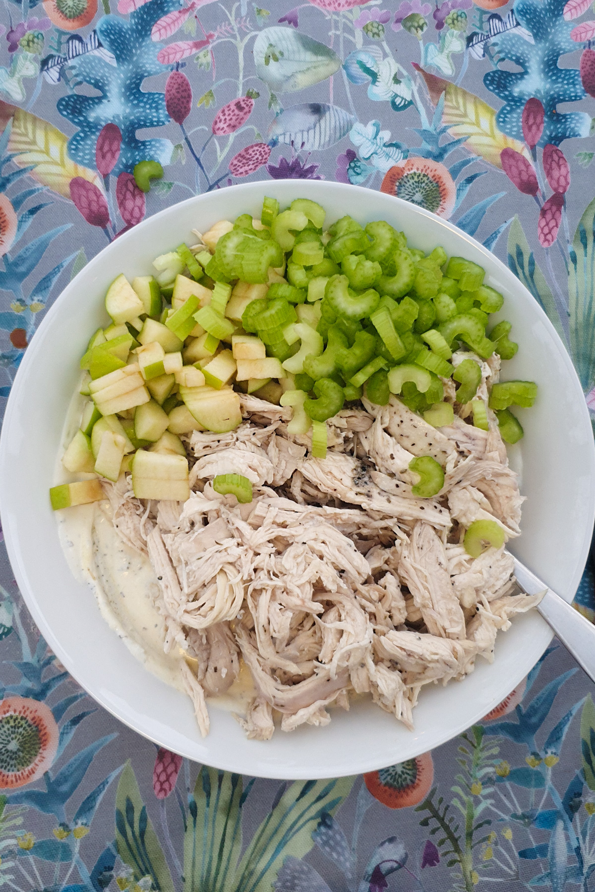 A white bowl containing shredded chicken, chopped celery, diced green apple, and creamy dressing, placed on a colorful floral tablecloth.