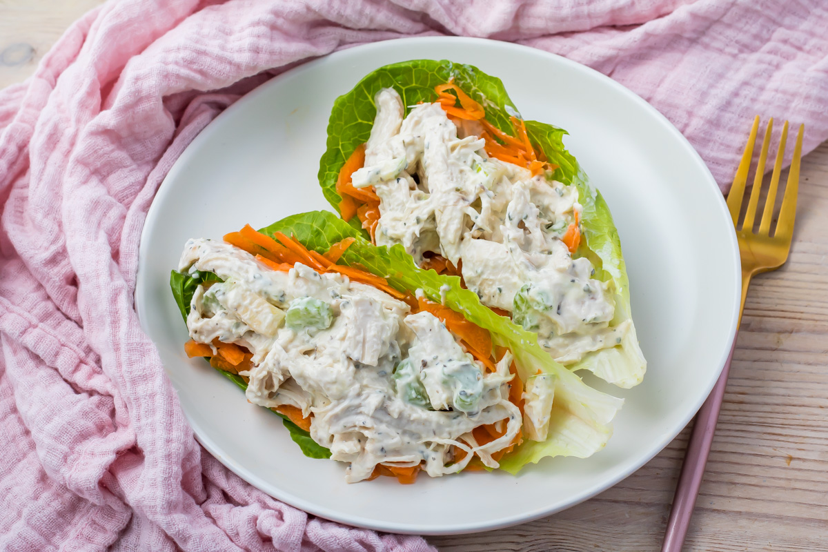Two high-protein chicken salad lettuce wraps topped with carrots, and creamy dressing are arranged on a white plate with a gold fork and a pink cloth nearby.