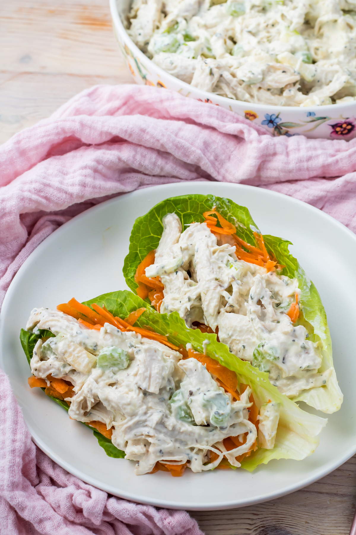 Two lettuce wraps filled with high-protein chicken salad and shredded carrots are served on a white plate. A pink cloth and a dish of extra chicken salad appear in the background.