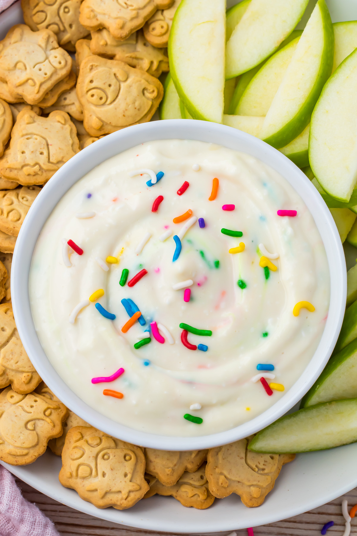 A bowl of creamy high protein funfetti dip with rainbow sprinkles, surrounded by green apple slices and animal-shaped crackers.