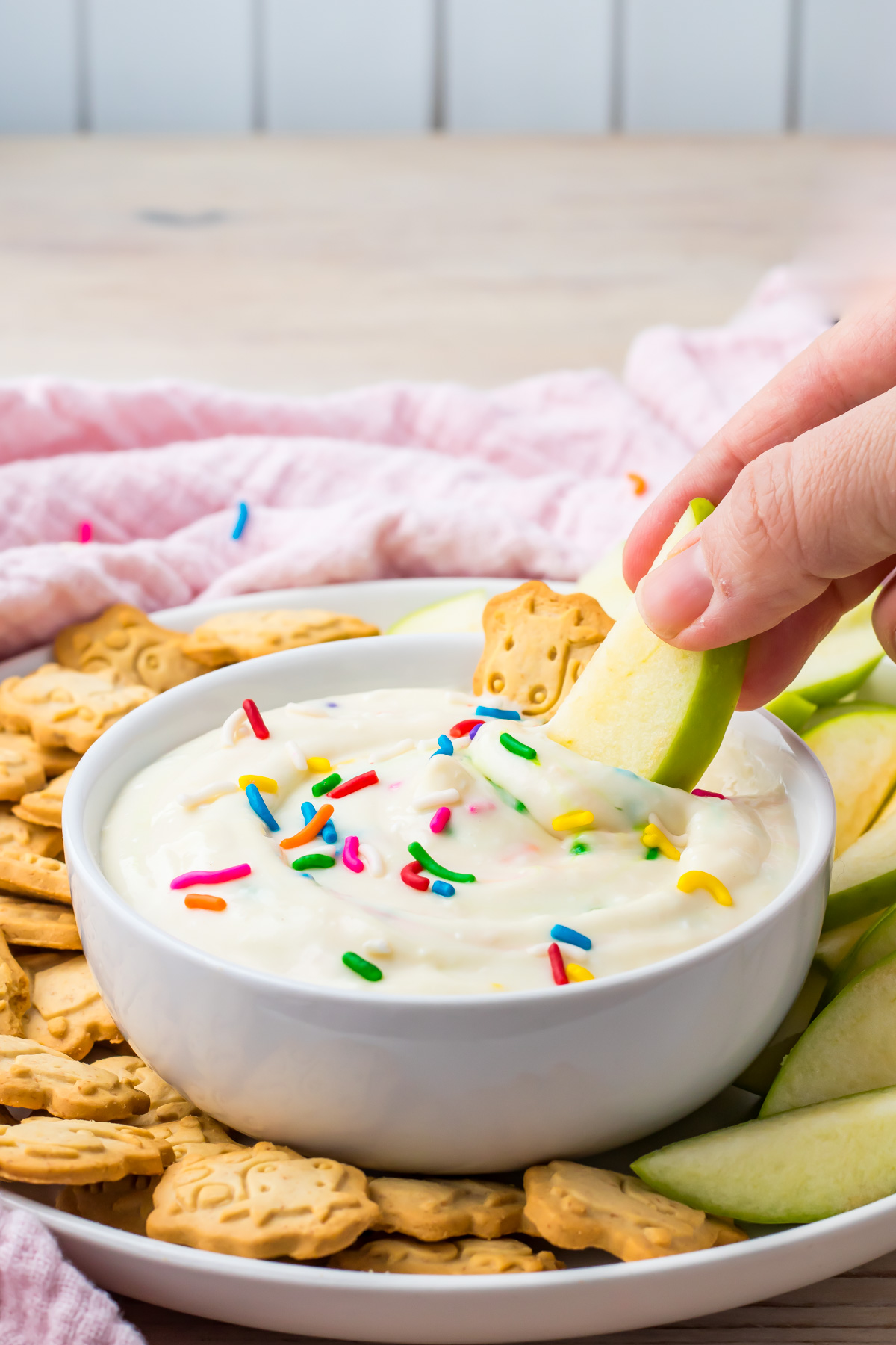 A hand dips a green apple slice into a bowl of high protein Funfetti Dip topped with colorful sprinkles, surrounded by crackers and more apple slices on a plate.