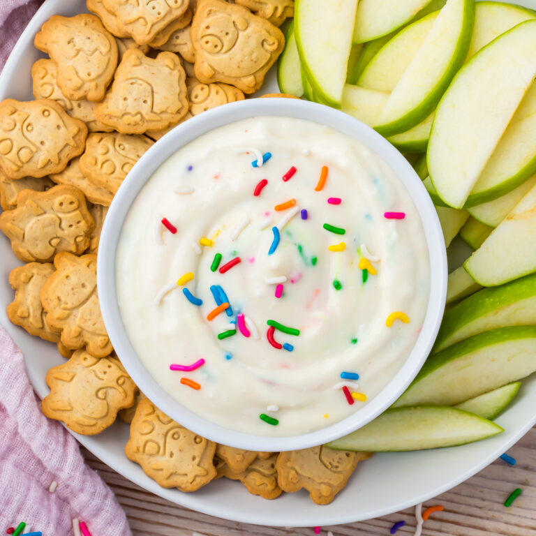 A bowl of high protein Funfetti Dip with colorful sprinkles is served alongside green apple slices and animal-shaped cookies on a white plate.