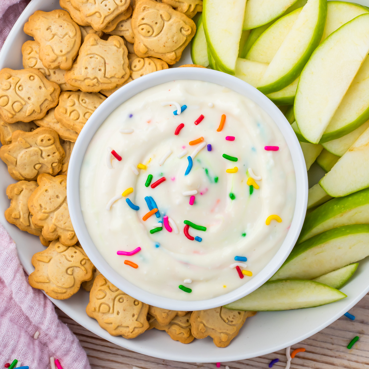 A bowl of high protein Funfetti Dip with colorful sprinkles is served alongside green apple slices and animal-shaped cookies on a white plate.