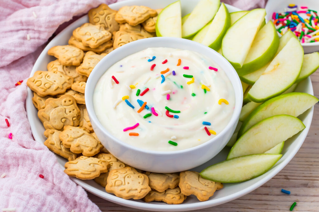 A white bowl of creamy high protein funfetti dip with colorful sprinkles sits on a white plate, surrounded by animal crackers and sliced green apples.