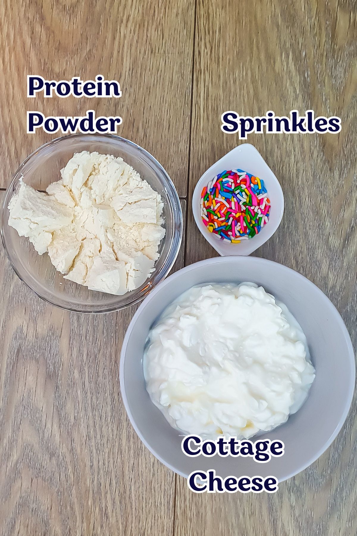 A bowl of protein powder, a small dish of colorful sprinkles, and cottage cheese are arranged on a wooden surface.