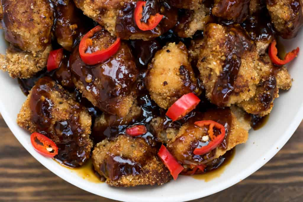 A bowl of breaded chicken pieces coated in a dark sauce and topped with sliced red chili peppers&mdash;perfect for those seeking high protein chicken recipes.