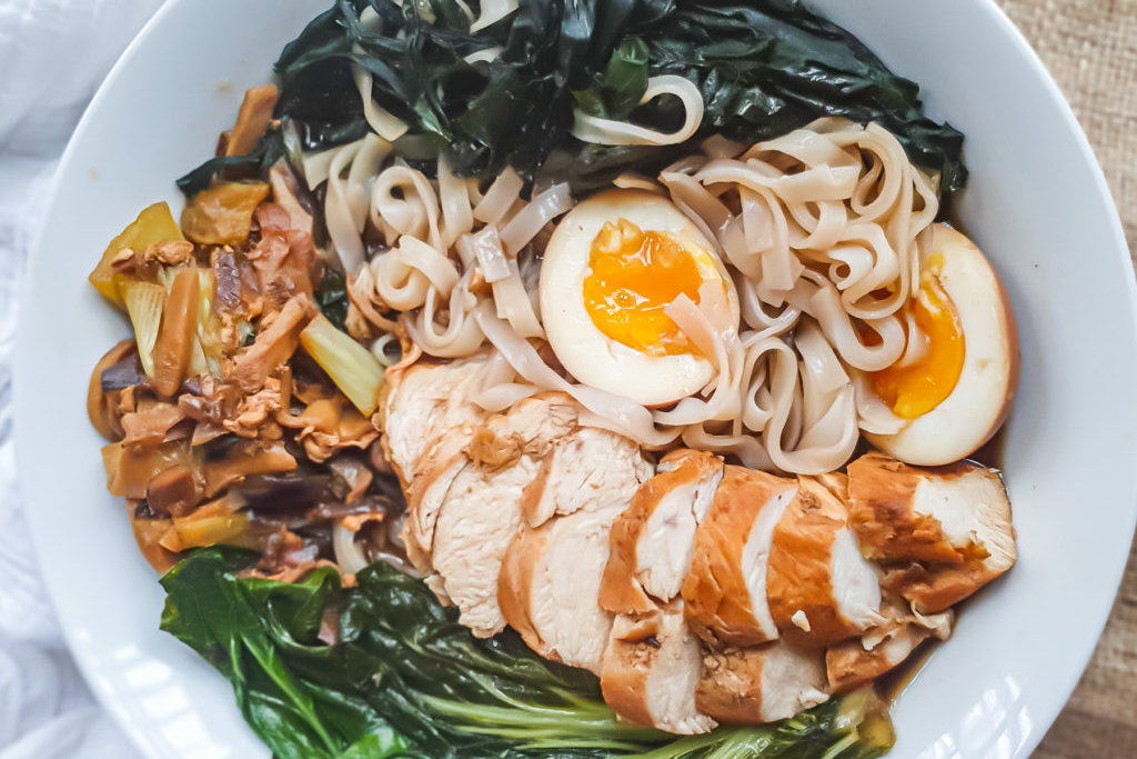 A bowl of noodles with high protein chicken breast, soft-boiled eggs, bok choy, seaweed, and assorted vegetables arranged neatly&mdash;perfect for those seeking flavorful high protein chicken recipes.