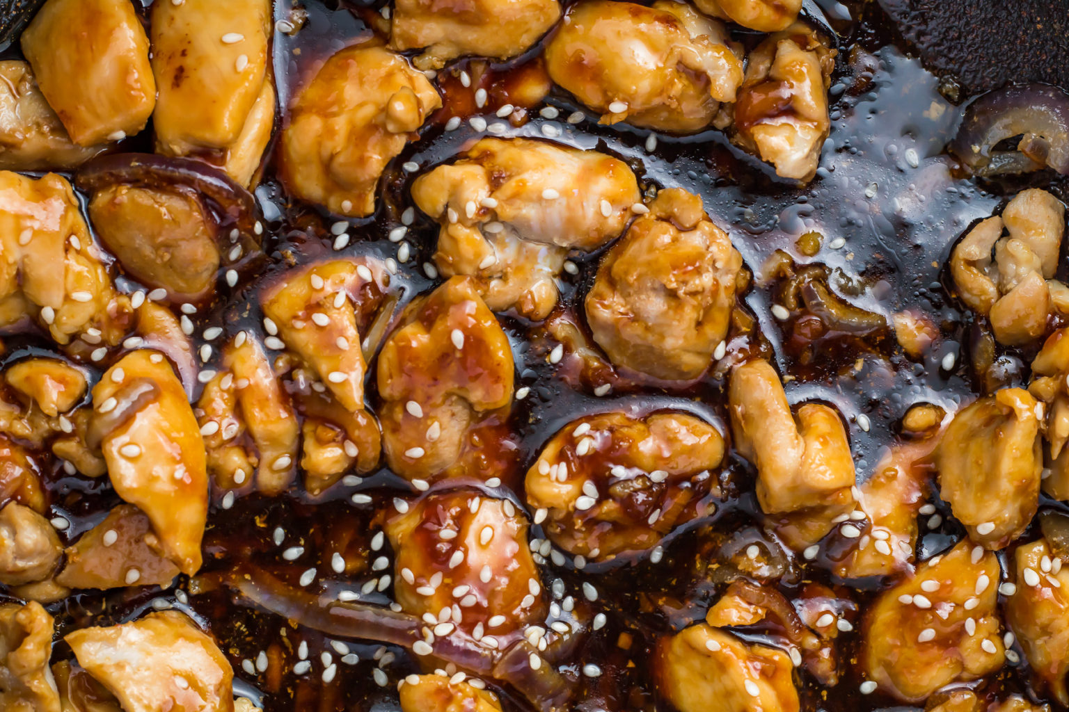 Close-up of cooked chicken pieces coated in a glossy brown sauce, garnished with white sesame seeds.