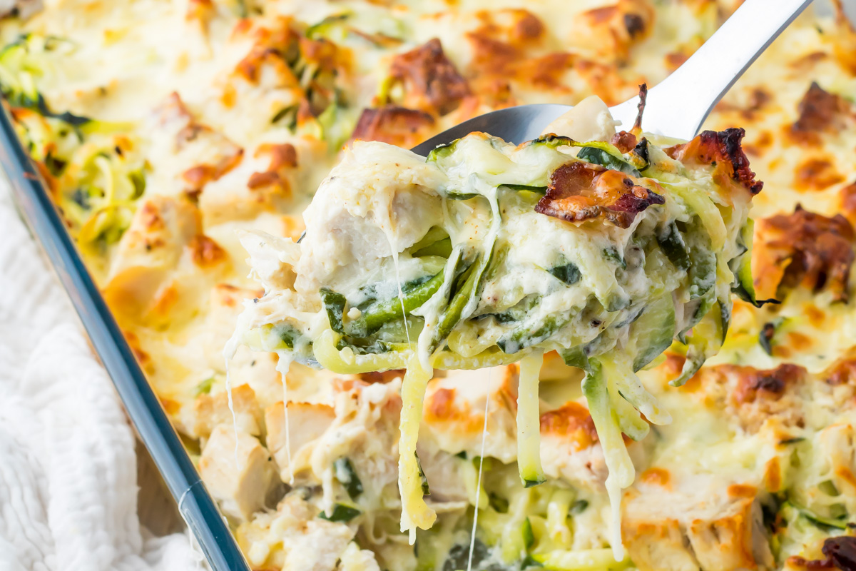 A close-up of a serving of cheesy baked keto chicken alfredo casserole, packed with shredded chicken, zucchini, spinach, and melted cheese being lifted from a baking dish.