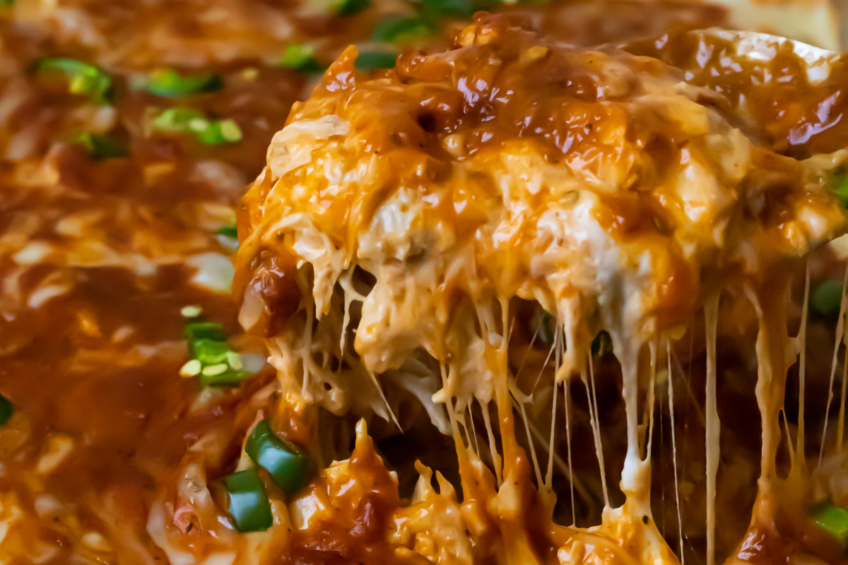 A close-up of cheesy keto chicken enchilada casserole being served, topped with melted cheese, red sauce, and chopped green onions.