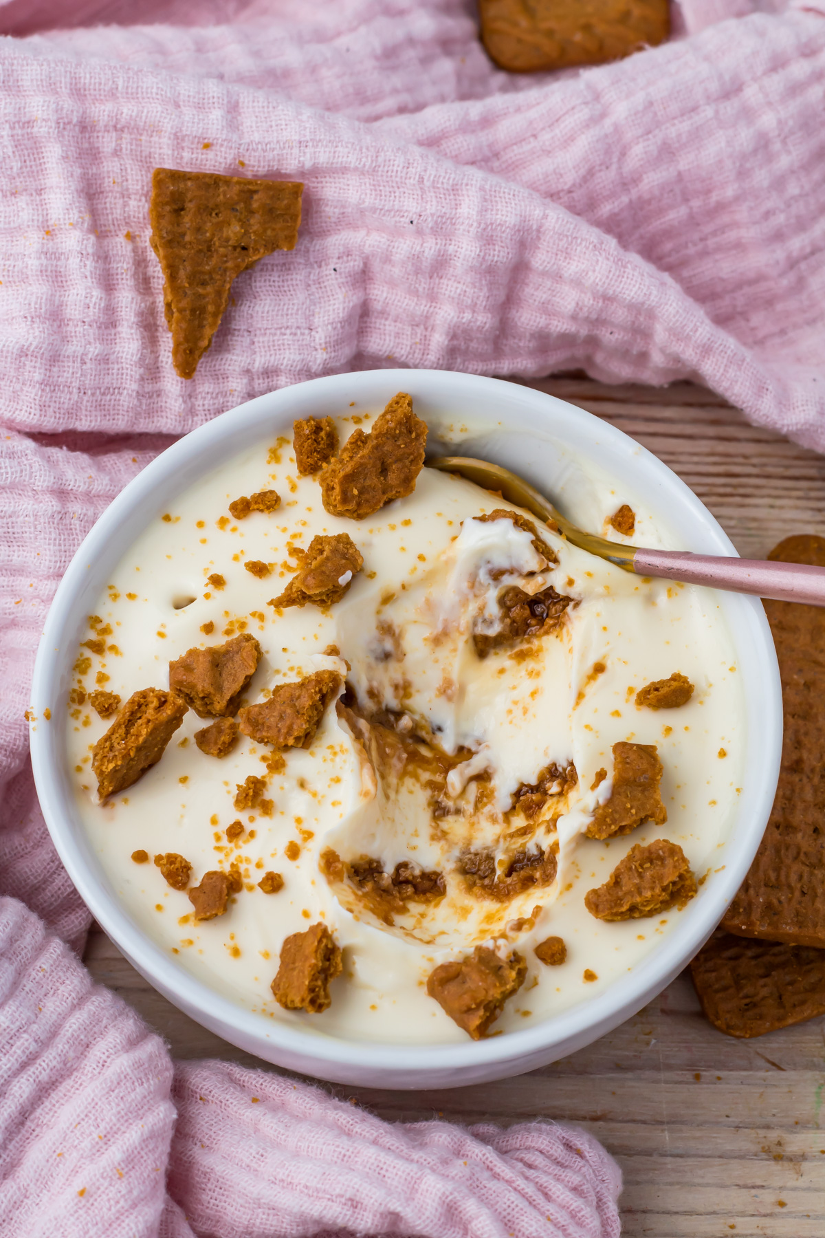 A bowl of creamy protein yogurt cheesecake topped with crumbled cookies, with a spoon inside, placed on a pink cloth and surrounded by cookie pieces.