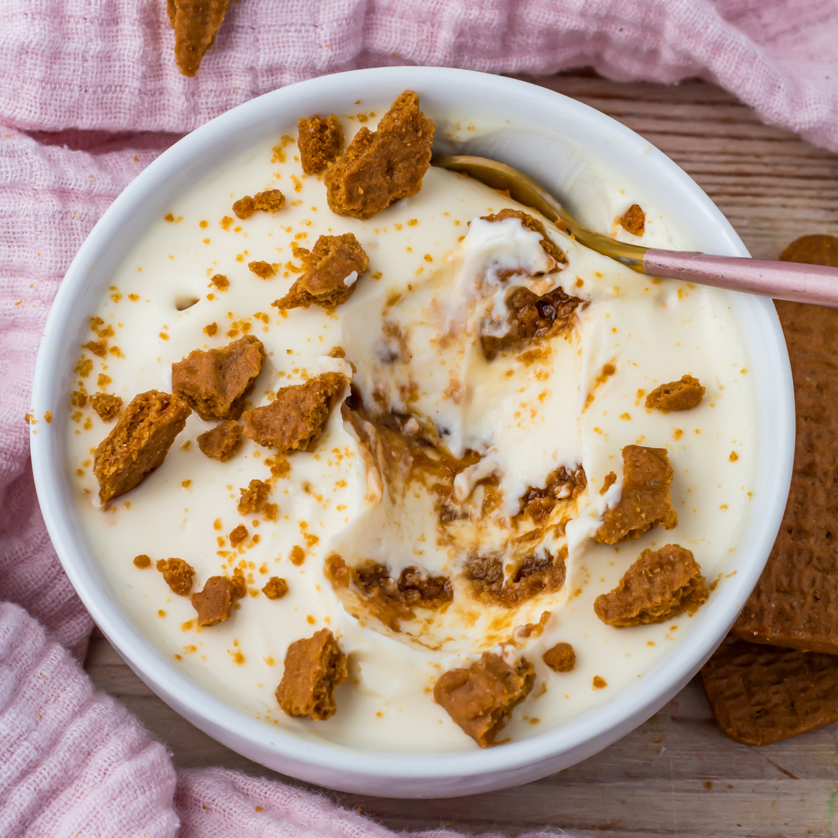 A bowl of creamy Protein Yogurt Cheesecake dessert topped with crumbled cookies, with a spoon resting inside.