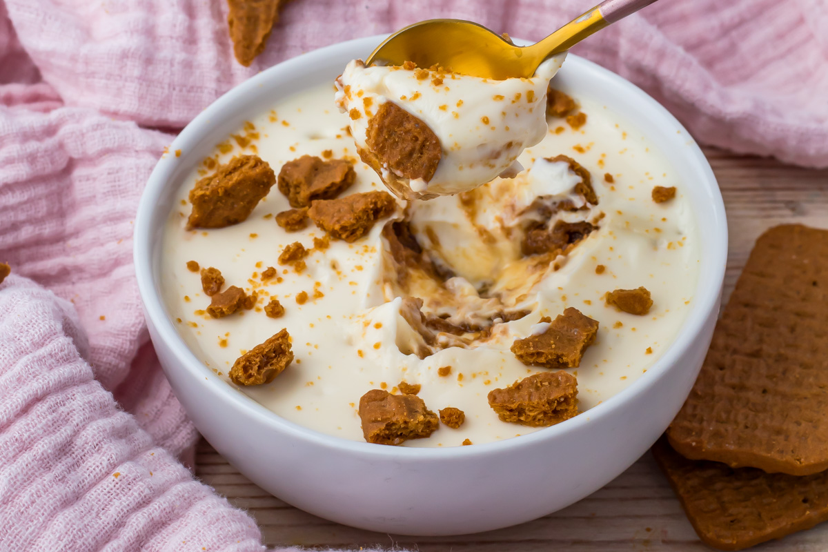 A bowl of creamy protein yogurt cheesecake dessert topped with crumbled cookies, with a spoonful being lifted out. Pink cloths and whole cookies are nearby.