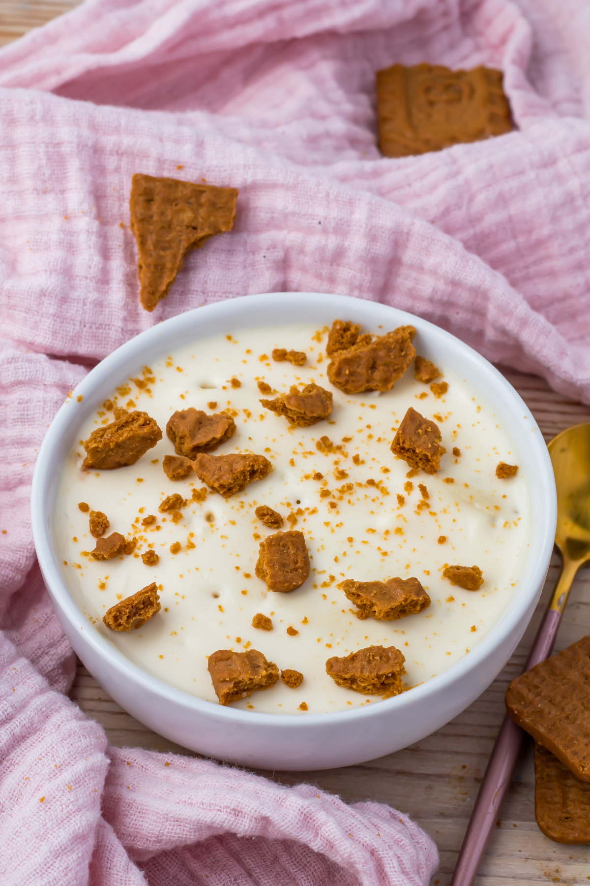 A white bowl filled with creamy protein yogurt cheesecake topped with crumbled brown cookies, placed on a pink textured cloth with extra cookie pieces nearby.