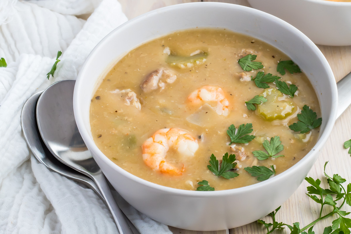 A bowl of thick seafood and chicken gumbo soup with shrimp, herbs, and vegetables, served with two metal spoons on a white cloth.