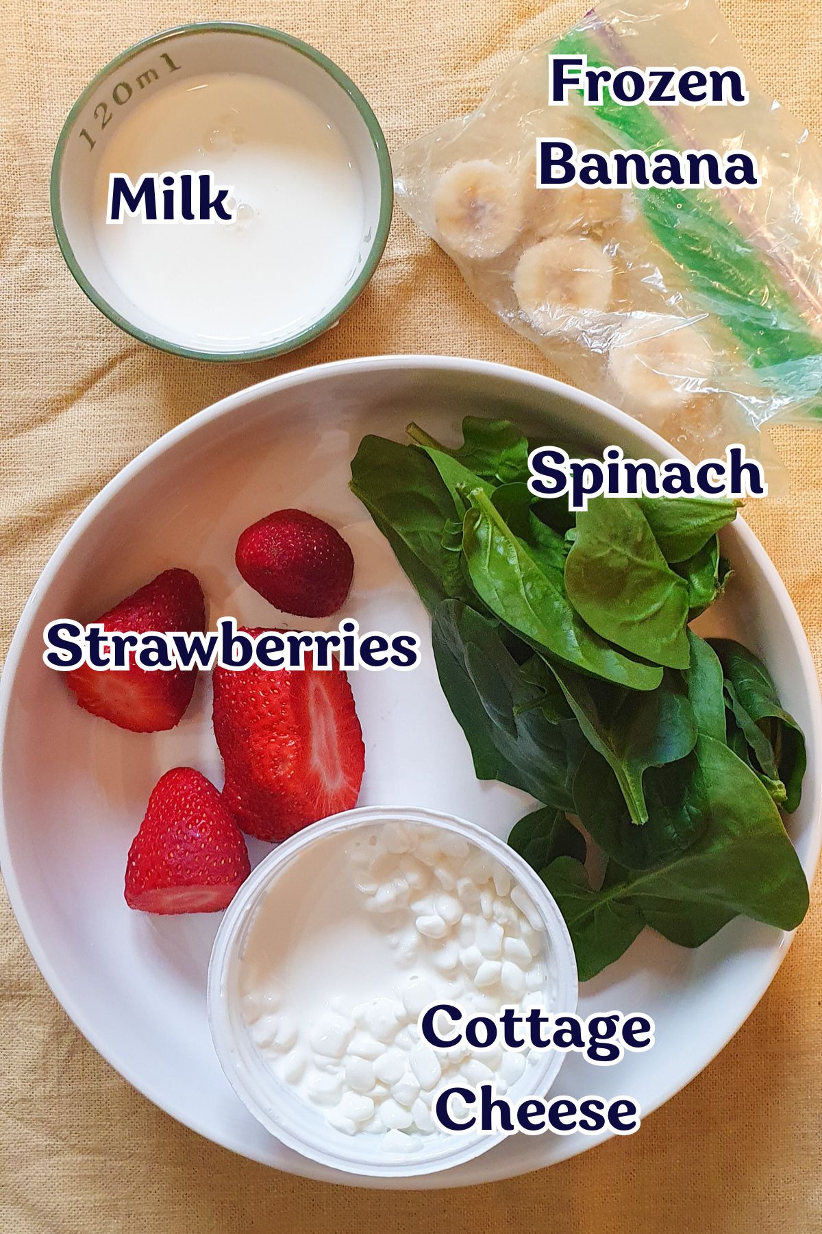 A bowl featuring strawberries, spinach, and a cottage cheese recipe, with separate servings of milk and frozen banana on a beige surface.