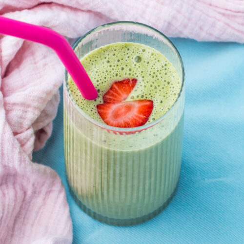 A glass of cottage cheese smoothie with a pink straw, topped with two strawberry slices, sits on a blue surface beside a pink cloth.