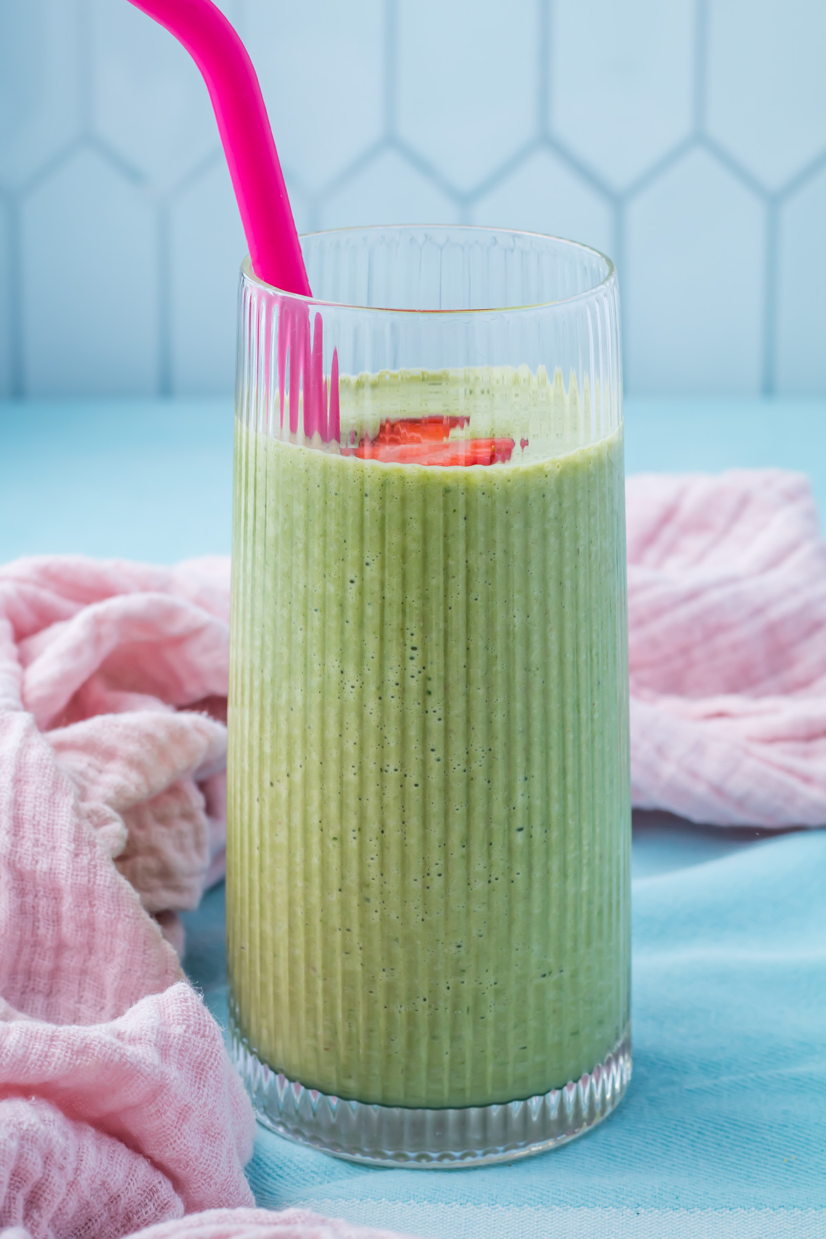 A tall glass of cottage cheese smoothie with a pink straw, garnished with sliced strawberries, placed on a blue surface next to a pink cloth.