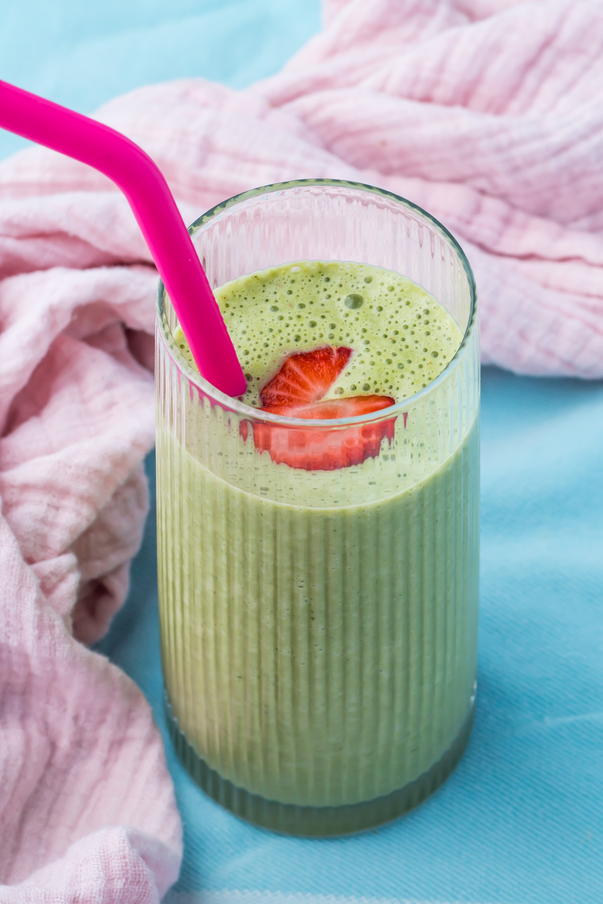 A glass of green cottage cheese smoothie with a pink straw, topped with sliced strawberries, sits on a light blue surface next to a pink cloth.