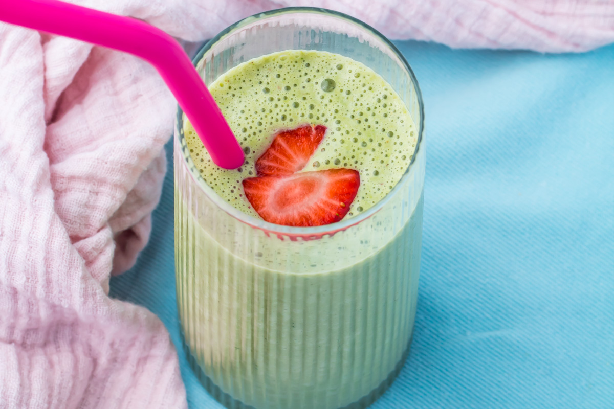 A glass of cottage cheese smoothie with two strawberry slices on top, served with a pink straw, placed on a blue surface next to a pink cloth.