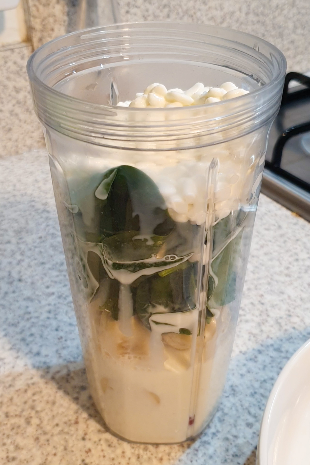 A blender cup filled with cottage cheese, spinach leaves, banana slices, and milk sits on a kitchen counter, ready to be blended into a nutritious cottage cheese smoothie.