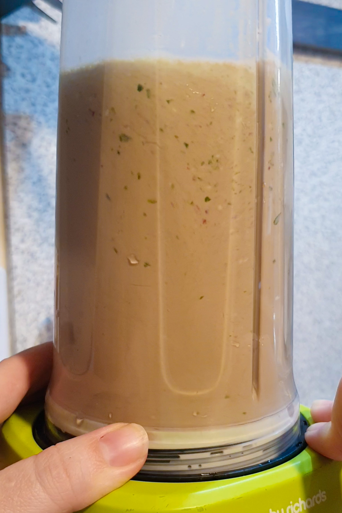 Close-up of a blender mixing a light brown cottage cheese smoothie with small green and red specks, held in place by two hands.