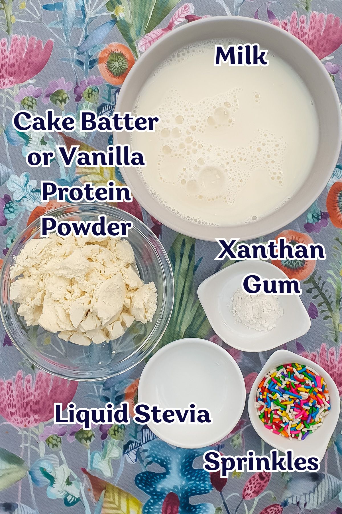 A bowl of milk, Cake Batter Protein powder, liquid stevia, xanthan gum, and colorful sprinkles sit on a floral surface—perfect for making delicious Protein Ice Cream.