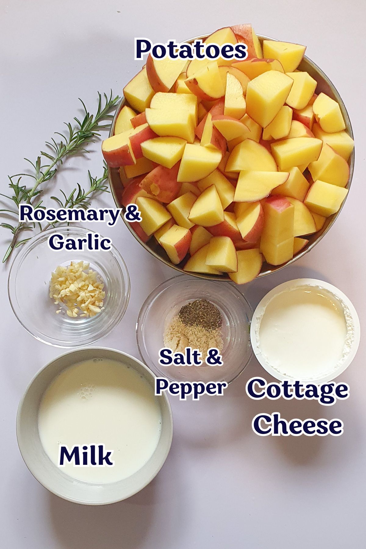 Chopped potatoes in a bowl, with bowls of cottage cheese, milk, salt and pepper, minced garlic, and a sprig of rosemary arranged on a white surface.
