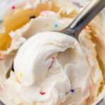 A close-up of a spoon scooping creamy white cake batter protein ice cream with colorful sprinkles from a glass jar.