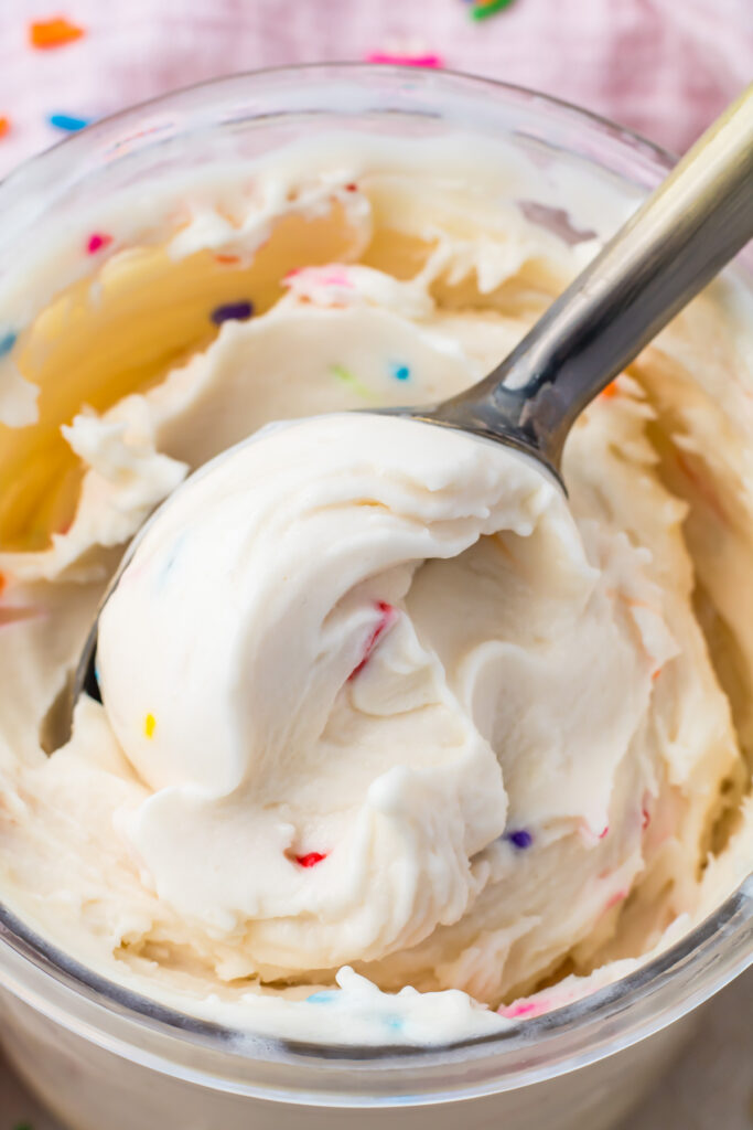 A close-up of a spoon scooping creamy white cake batter protein ice cream with colorful sprinkles from a glass jar.