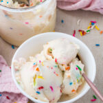 A white bowl with scoops of cake batter vanilla ice cream containing colorful rainbow sprinkles, next to a container of more protein ice cream and a pink cloth, with sprinkles scattered on the table.