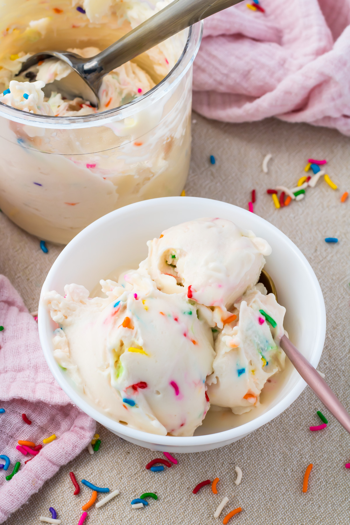 A white bowl with scoops of cake batter ice cream containing colorful rainbow sprinkles, next to a container of more protein ice cream and a pink cloth, with sprinkles scattered on the table.