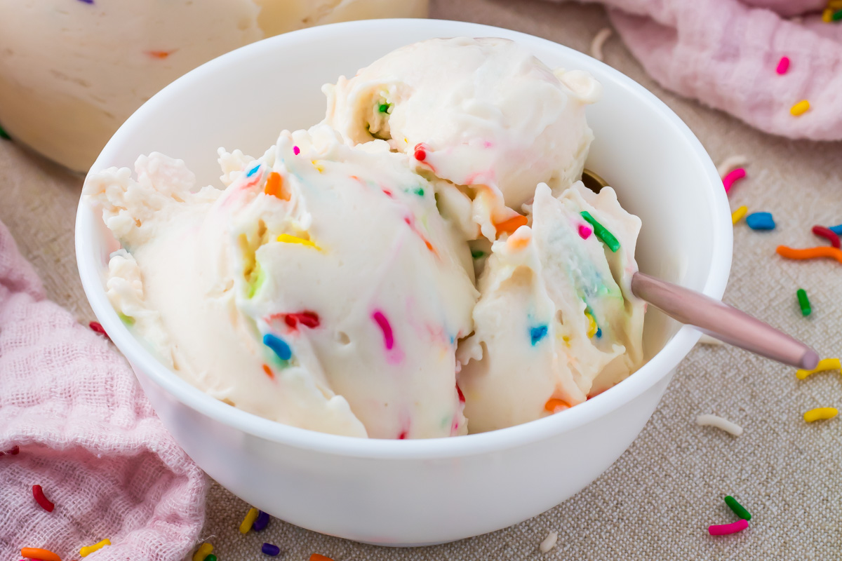 A white bowl filled with Cake Batter Protein ice cream mixed with colorful sprinkles, with a spoon resting on the side and scattered sprinkles nearby.