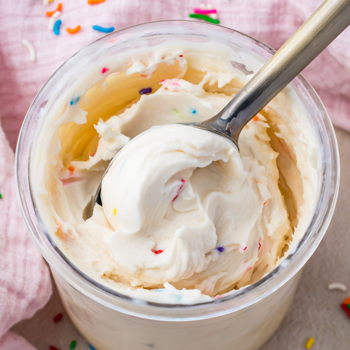 A spoon in a cup of Cake Batter Protein Ice Cream with white frosting and colorful sprinkles, set on a pink cloth.