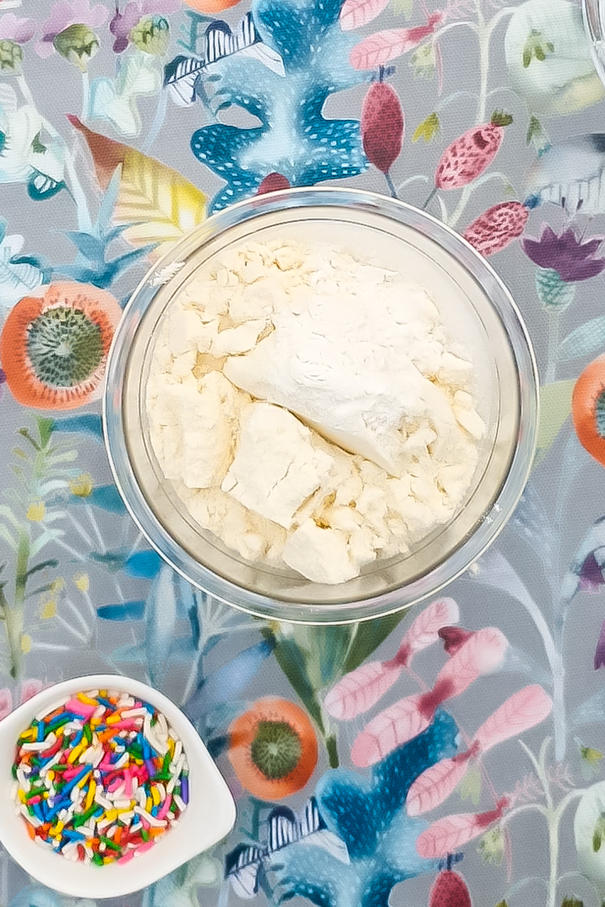 A creami pint containing an ice cream mixture sits on a colorful floral tablecloth, next to a small white dish filled with rainbow sprinkles.