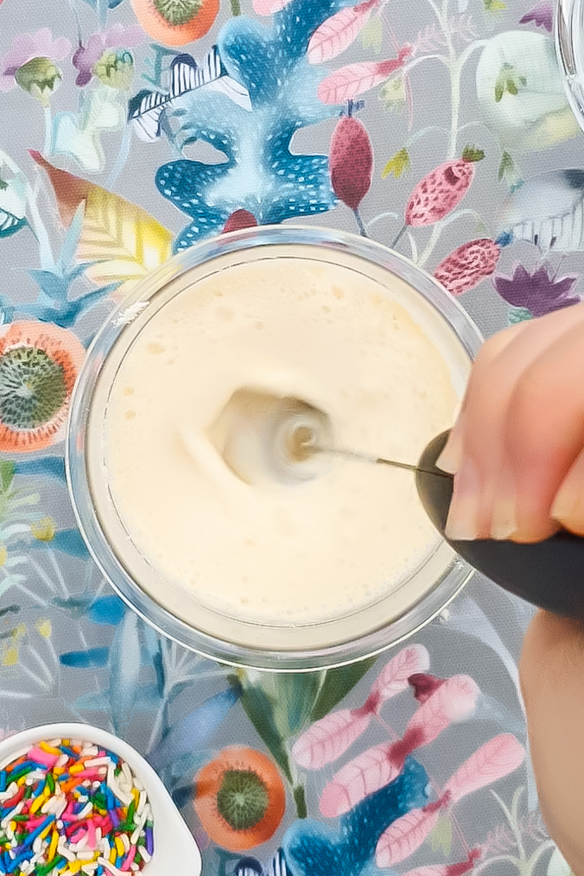 A hand uses a handheld milk frother to mix a light ice cream mixture in a ninja creami pint, with a bowl of colorful sprinkles and a floral-patterned surface underneath.
