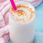 A glass of creamy Cinnamon Roll Protein Shake with a pink straw, topped with a swirl of ground cinnamon, sits on a light blue surface next to a pink textured cloth.