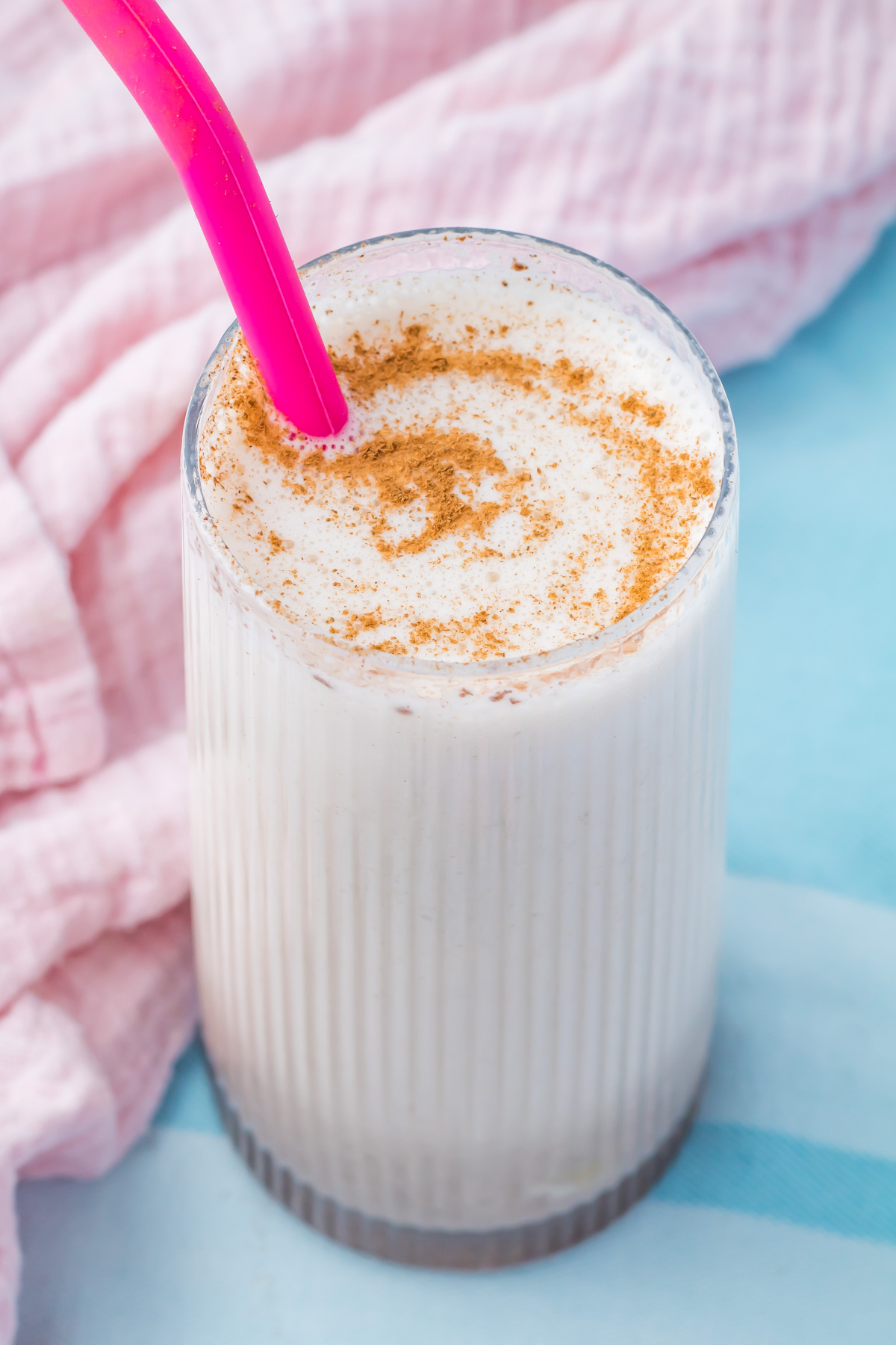 A glass of creamy Cinnamon Roll Protein Shake with a pink straw, topped with a swirl of ground cinnamon, sits on a light blue surface next to a pink textured cloth.