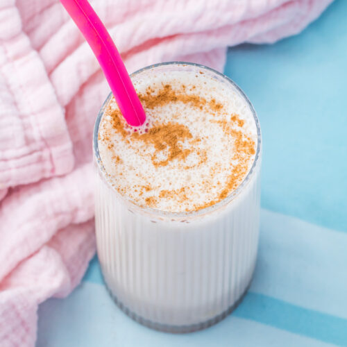 A glass of creamy Cinnamon Roll Protein Shake with a pink straw, topped with a sprinkle of cinnamon, sits on a blue surface next to a pink textured cloth.