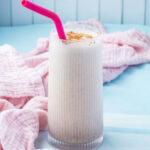 A tall glass of creamy Cinnamon Roll Protein Shake with a pink straw and sprinkled cinnamon on top, placed on a light blue surface with a pink cloth.