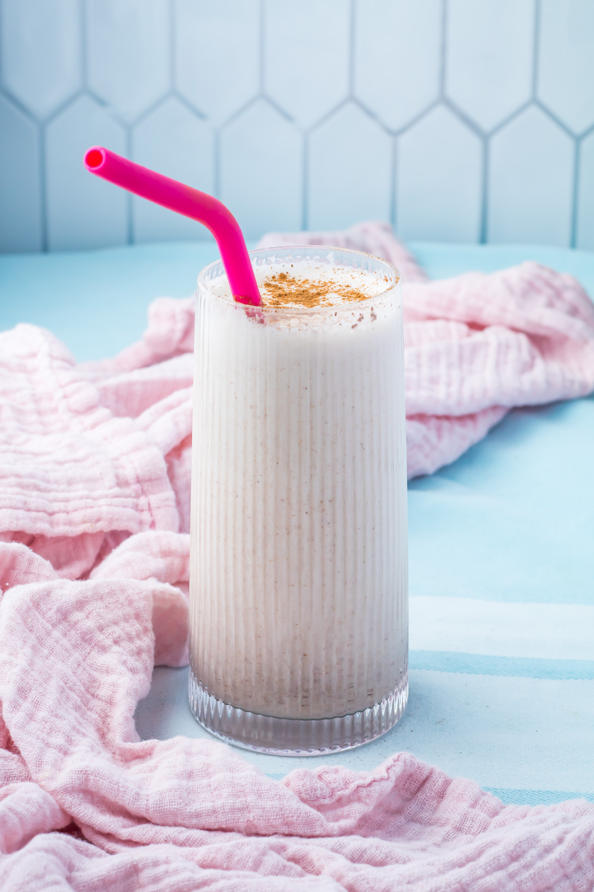 A tall glass of creamy Cinnamon Roll Protein Shake with a pink straw and sprinkled cinnamon on top, placed on a light blue surface with a pink cloth.