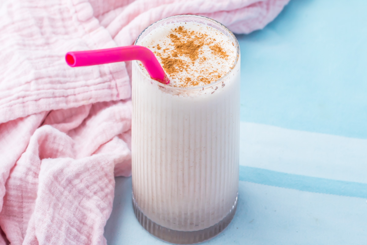 A glass of Cinnamon Roll Protein Shake with a pink straw, topped with cinnamon, sits on a light blue surface next to a pink cloth&mdash;reminiscent of the flavors in a refreshing cinnamon roll protein shake.
